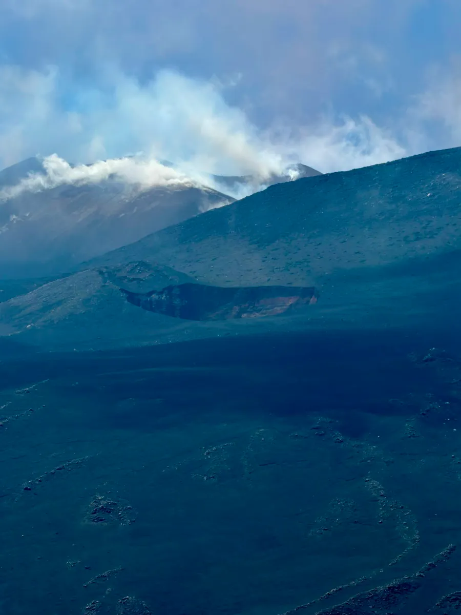 Pit crater riconoscibile da lontano come un taglio scuro sul fianco dell'Etna, con i crateri sommitali che degassano sullo sfondo